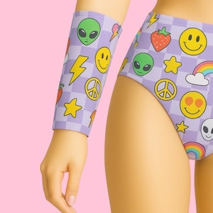 May include: A lilac-colored, checkered pattern arm sleeve and high-waisted bottoms. The design features various colorful graphics, including smiley faces, alien heads, strawberries, stars, peace signs, lightning bolts, and rainbows.