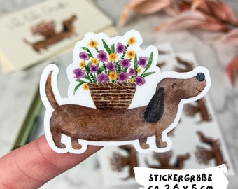 May include: A brown dachshund carrying a basket of purple, yellow, and white flowers. The sticker is approximately 3.6 inches by 5 inches. "STICKERGRÖSSE CA. 3,6 X 5 CM" is written in black text on a white background.