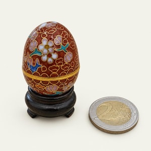 Op de afbeelding: Een kleine eivormige cloisonné houder met een dieprode achtergrond, versierd met witte en lichtpaarse bloemmotieven en gouddraadcontouren. Het staat op een donkere houten standaard naast een munt van 2 euro voor schaalvergelijking.