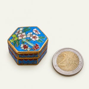 Pode incluir: Uma caixa hexagonal de cloisonné com fundo azul brilhante, apresentando padrões florais brancos e vermelhos com folhas verdes e bordas de metal dourado. Uma moeda de 2 euros está ao lado da caixa para indicar a sua pequena escala.