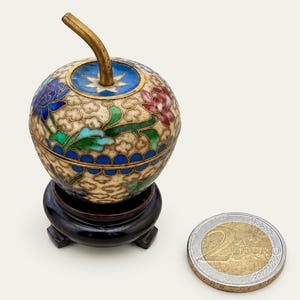 Op de afbeelding: Een kleine, ronde, appelvormige cloisonné houder met een koperen steeltje, voorzien van bloemmotieven in blauw, roze en groen op een crèmekleurige achtergrond. Het staat op een donkere houten standaard naast een 2 euro munt voor schaal.