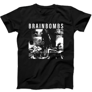 May include: A black short-sleeve t-shirt featuring a white graphic print on the chest. The print displays the text BRAINBOMBS in a bold sans-serif font above a high-contrast, grainy black and white photograph of a room with figures.