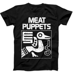 May include: A black short-sleeve t-shirt featuring a white distressed graphic print. The text MEAT PUPPETS appears in a bold, weathered font above a square frame containing a stylized, abstract bird-like figure with geometric patterns.