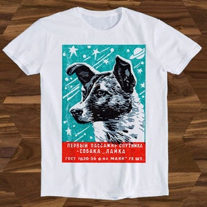 Könnte beinhalten: Weißes T-Shirt mit einer Vintage-Illustration eines Hundes im Weltraum. Die Illustration ist von einem türkisfarbenen Rand und Sternen umgeben. Der Text lautet "ПЕРВЫЙ ПАССАЖИР СПУТНИКА - СОБАКА...ЛАЙКА ГОСТ 1820-56 Ф-КА "МАЙК" 75 ШТ."