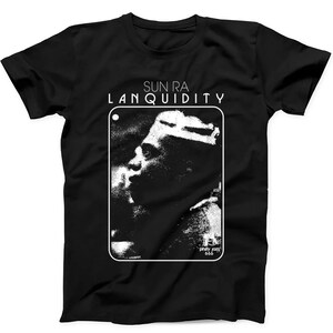 Puede incluir: Una camiseta negra de manga corta con un estampado gráfico blanco de Sun Ra. El diseño incluye el texto SUN RA LANQUIDITY en la parte superior y Philly Jazz 666 en la parte inferior derecha, dentro de un marco rectangular blanco.