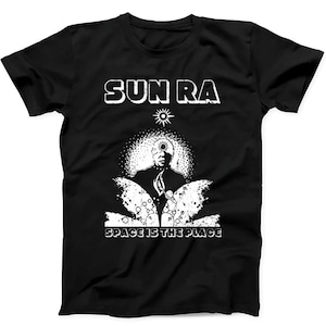 Puede incluir: Una camiseta negra de manga corta con un estampado gráfico en blanco. El diseño incluye el texto SUN RA en la parte superior y SPACE IS THE PLACE en la inferior. La imagen central muestra una figura con cabeza solar y elementos cósmicos.