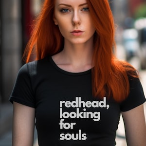 May include: A woman with long, bright red hair is wearing a black t-shirt with the text "redhead, looking for souls" printed on it.