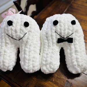 Puede incluir: Dos peluches blancos en forma de U con ojos negros de botón y sonrisas cosidas. Uno tiene un lazo rosa, el otro una pajarita negra. Los juguetes están hechos de un material suave y texturizado, perfectos para una guardería o como regalo.