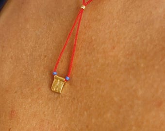 May include: A delicate necklace featuring a red cord with a small, textured gold-colored pendant. The pendant is rectangular with a hammered finish, and is accented with small blue beads. The necklace is simple and elegant.