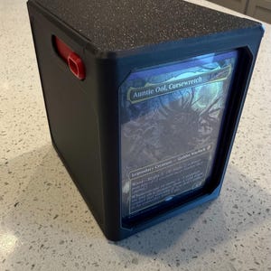 Könnte beinhalten: Eine schwarze, rechteckige Kartenbox mit strukturierter Oberseite und einem roten seitlichen Verschluss. Die Vorderseite verfügt über ein transparentes Sichtfenster, in dem eine Magic: The Gathering-Sammelkarte namens Auntie Ool, Cursewretch liegt.