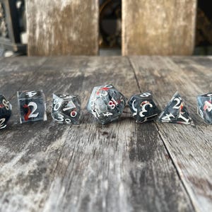 May include: A set of seven polyhedral dice with a black and clear design, featuring white numbers and red accents. The dice are arranged on a weathered wooden surface, showcasing various geometric shapes.