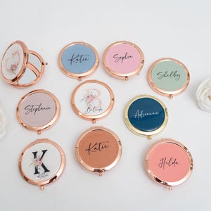 May include: A collection of ten round, compact mirrors with rose gold trim. Each mirror has a different design, including floral patterns, initials, and names. The mirrors are arranged in a circle on a white surface.