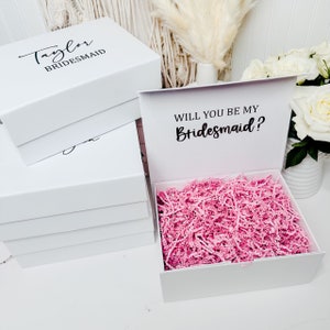 May include: White gift box with a pink tissue paper filler. The lid of the box has the text "Will you be my Bridesmaid?" in black script font. The box is closed and stacked on top of other white boxes with the text "Taylor Bridesmaid" on the top box.