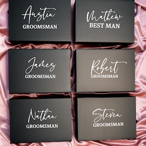 May include: Six black gift boxes with white text on a pink satin surface. The boxes are labeled with names and titles: Austin, James, Nathan (Groomsman), Matthew (Best Man), Robert, and Steven (Groomsman).