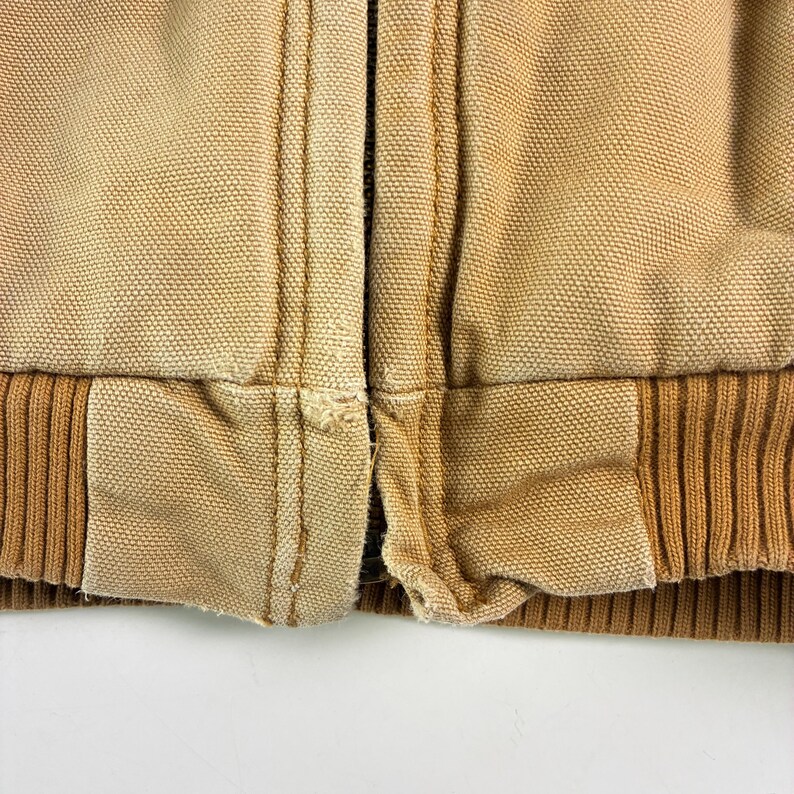 May include: Close-up of a tan work jacket. The jacket features a ribbed brown hem and cuffs, with a tan canvas body. Visible stitching and a zipper are present. The fabric appears worn, with some fraying.