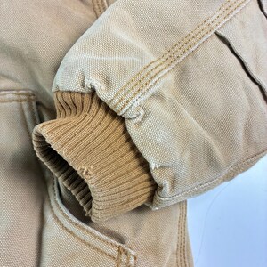 May include: Close-up of a tan work jacket sleeve. The sleeve features a ribbed knit cuff in a lighter tan color. The jacket fabric is a durable canvas with visible stitching in a contrasting tan thread.