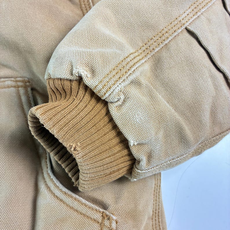May include: Close-up of a tan work jacket sleeve. The sleeve features a ribbed knit cuff in a lighter tan color. The jacket fabric is a durable canvas with visible stitching in a contrasting tan thread.