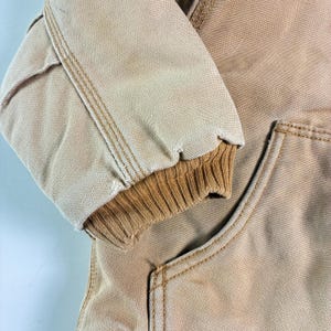 May include: Close-up of a tan work jacket sleeve with a ribbed cuff. The jacket has visible stitching in a contrasting tan color. The fabric appears to be a durable, heavy-duty material, with a pocket visible on the lower right.