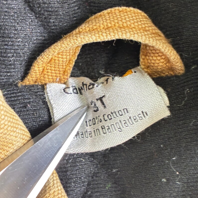 May include: Close-up of a Carhartt clothing tag. The tag is white with black text that reads "Carhartt", "3T", "100% Cotton", and "Made in Bangladesh". The tag is being cut with scissors. The background is black.