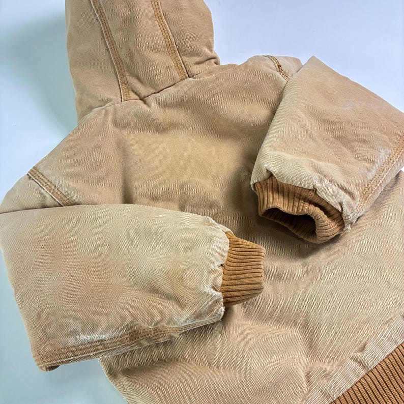 May include: A tan hooded jacket with ribbed cuffs and hem. The jacket has a worn appearance, with visible stitching and a sturdy, durable design. The hood is attached, and the sleeves are long. The jacket is made of a heavy-duty material.