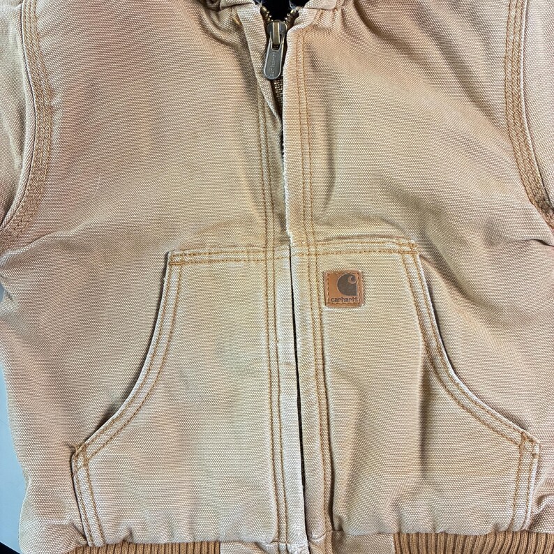 May include: Close-up of a tan Carhartt jacket with a hood and a zipper closure. The jacket has two front pockets and a small Carhartt logo patch. The stitching is visible, and the jacket appears to be made of a durable material.