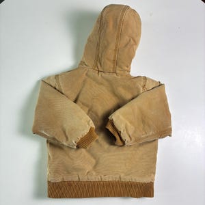 May include: A tan hooded jacket with a brown ribbed hem and cuffs. The jacket has a full hood and is made of a durable, workwear-style fabric. The jacket is designed for warmth and protection from the elements.