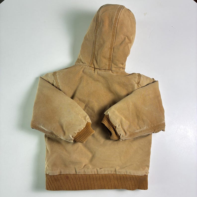 May include: A tan hooded jacket with a brown ribbed hem and cuffs. The jacket has a full hood and is made of a durable, workwear-style fabric. The jacket is designed for warmth and protection from the elements.