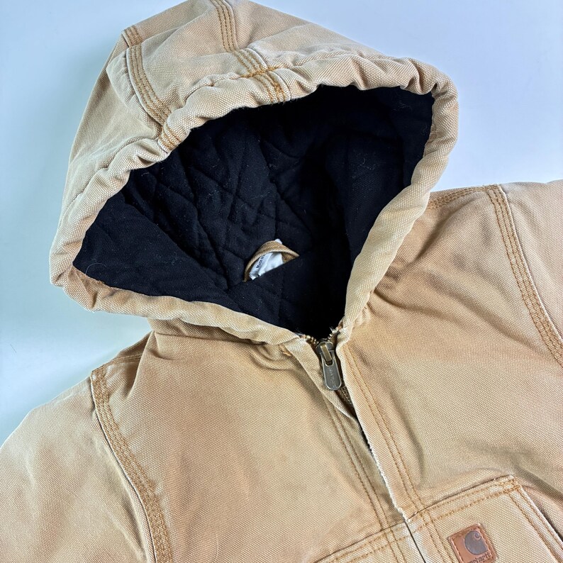 May include: Close-up of a tan hooded jacket with a black quilted lining. The jacket features a full zipper closure and visible stitching details. A small, brown logo patch is on the lower right side.