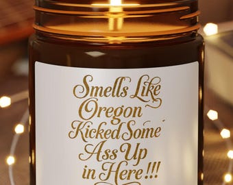 May include: A lit brown glass candle with a white label that reads "Smells Like Oregon Kicked Some Ass Up in Here!!!" The candle sits on a small wooden coaster, surrounded by string lights. A guitar is visible in the background.
