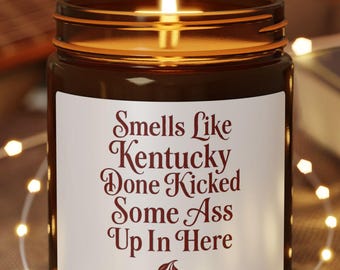 May include: A lit brown glass candle jar with a white label that reads "Smells Like Kentucky Done Kicked Some Ass Up In Here." The candle sits on a small wooden coaster, with a string of lights and a guitar in the background.