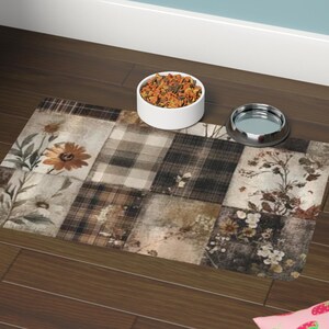 May include: A rectangular pet feeding mat with a patchwork design in shades of brown, beige, and cream. The mat features floral and plaid patterns. A white ceramic bowl and a silver bowl sit on the mat.
