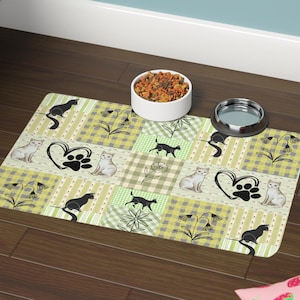May include: A rectangular pet food mat with a patchwork design in shades of green, white, and tan. The mat features cat silhouettes, paw prints, and floral patterns. A white ceramic bowl and a stainless steel bowl sit on the mat.