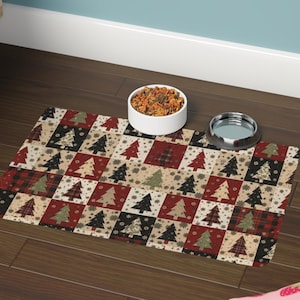 May include: A rectangular pet food mat with a patchwork design featuring Christmas trees in red, black, and beige squares. A white ceramic bowl filled with pet food and a stainless steel bowl sit on the mat.