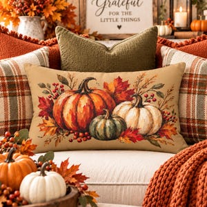 May include: A rectangular throw pillow featuring a printed illustration of three pumpkins in orange, green, and white, surrounded by autumn leaves and red berries on a tan background, positioned in front of several solid and plaid decorative pillows.