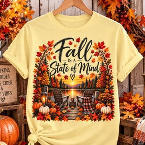 May include: A pale yellow t-shirt featuring a graphic of two wooden chairs on a dock overlooking a lake at sunset. The design includes autumn leaves, pumpkins, a lantern, and the text "Fall IS A State of Mind" with a small heart symbol.