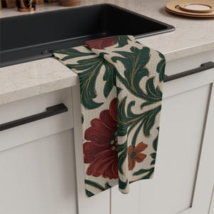 May include: A decorative kitchen towel featuring a floral pattern with large red flowers and green leaves on a cream background. The towel is draped over a black sink and white cabinet.