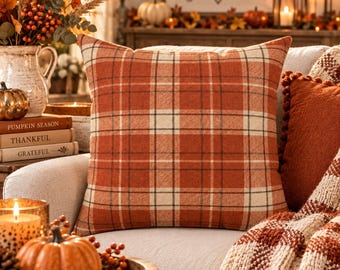 May include: A square throw pillow featuring a plaid pattern in shades of burnt orange, cream, and dark brown. The fabric has a visible woven texture, and the pillow is positioned on a light-colored upholstered chair with other seasonal decor.