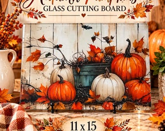 May include: A rectangular glass cutting board featuring a printed illustration of orange and white pumpkins, a dark bucket, and autumn leaves against a white wooden background. The text reads Autumn Pumpkins GLASS CUTTING BOARD 11 x 15 INCHES.