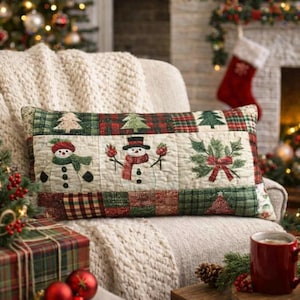 Könnte beinhalten: Rechteckiges Weihnachtskissen mit Patchwork-Design. Das Kissen zeigt gestickte Schneemänner, Weihnachtsbäume und einen Stechpalmenzweig mit roter Schleife. Das Farbschema umfasst Rot, Grün und Creme.