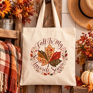 May include: A cream-colored canvas tote bag featuring a central graphic of a large, multi-colored maple leaf surrounded by a circular floral wreath. The text "Fall Is My Favorite Season" is printed in a decorative script font around the leaf.