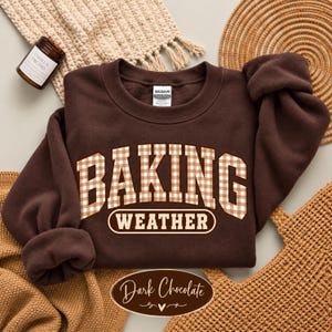 May include: A dark brown crewneck sweatshirt featuring the text BAKING WEATHER in a white and tan gingham pattern. The word BAKING is in a large collegiate font, while WEATHER is enclosed in a white oval outline. A Gildan brand tag is visible.