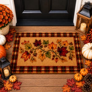 May include: A rectangular doormat featuring a central tan field decorated with a sprawling vine of colorful autumn maple leaves in shades of red, orange, and green, framed by a dark red, green, and gold plaid border pattern.