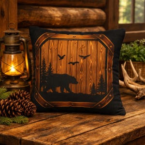 May include: Black square pillow with a wooden frame design. The center features a silhouette of a bear, trees, and birds against a wood-grain background. The pillow is on a wooden surface, with a lit lantern and pine cones in the background.