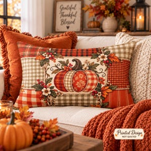 Könnte beinhalten: Ein rechteckiges Zierkissen mit einer zentralen Kürbisillustration, umgeben von Herbstblättern und Beeren. Das Design zeigt ein Patchwork-Muster aus roten, orangen und beigen Karos, Vichy-Mustern und gestreiften Stoffblöcken auf cremefarbenem Grund.