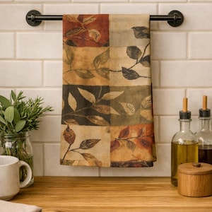 May include: A decorative kitchen towel with a leaf pattern in shades of brown, beige, and green hangs on a black metal bar. The towel is displayed in a kitchen setting with a wooden countertop and white tile backsplash.