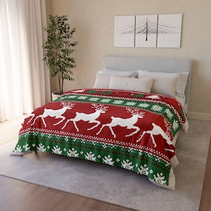 May include: A red and green fleece blanket featuring a repeating white reindeer pattern and snowflake motifs. The blanket is draped over a bed with white pillows and a grey headboard, showcasing a festive design with horizontal bands.