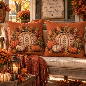 May include: Two square throw pillows with an orange background featuring a central large white pumpkin surrounded by smaller orange and green pumpkins, autumn leaves, and berries. A sign above reads PUMPKINS HAYRIDES FALLING LEAVES Cozy Days.