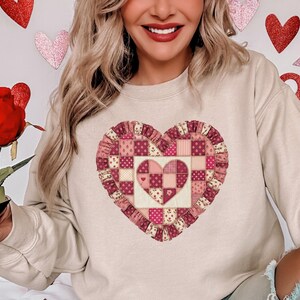 May include: Beige sweatshirt featuring a large heart design. The heart is a patchwork of pink and red fabrics, with a ruffled border. The design includes floral and polka dot patterns. The sweatshirt is a neutral color.