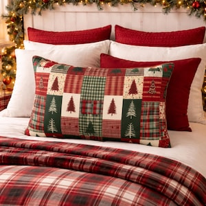 May include: A bed is adorned with red and white pillows, a plaid blanket, and a decorative pillow with a patchwork design. The patchwork pillow features Christmas trees and plaid patterns in red and green. A garland with lights and ornaments decorates the headboard.