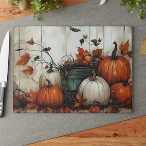May include: Rectangular glass cutting board featuring a fall harvest scene. The design includes orange and white pumpkins, fall leaves, and a rustic wooden background. A knife is visible on the left side of the board.